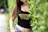 A person with long brown hair is wearing a black tank top with a 'Real Poker' logo in yellow and white, standing partially behind green foliage. They are also wearing blue jeans, showcasing a casual look.
