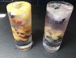 Two glasses filled with a clear fizzy beverage and ice cubes are placed on a dark surface. The glass on the left contains slices of yellow and red fruit, while the glass on the right has purple and light-green fruit pieces.