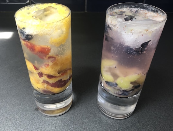 Two glasses filled with a clear fizzy beverage and ice cubes are placed on a dark surface. The glass on the left contains slices of yellow and red fruit, while the glass on the right has purple and light-green fruit pieces.