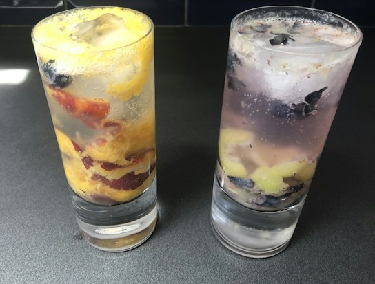 Two glasses filled with a clear fizzy beverage and ice cubes are placed on a dark surface. The glass on the left contains slices of yellow and red fruit, while the glass on the right has purple and light-green fruit pieces.