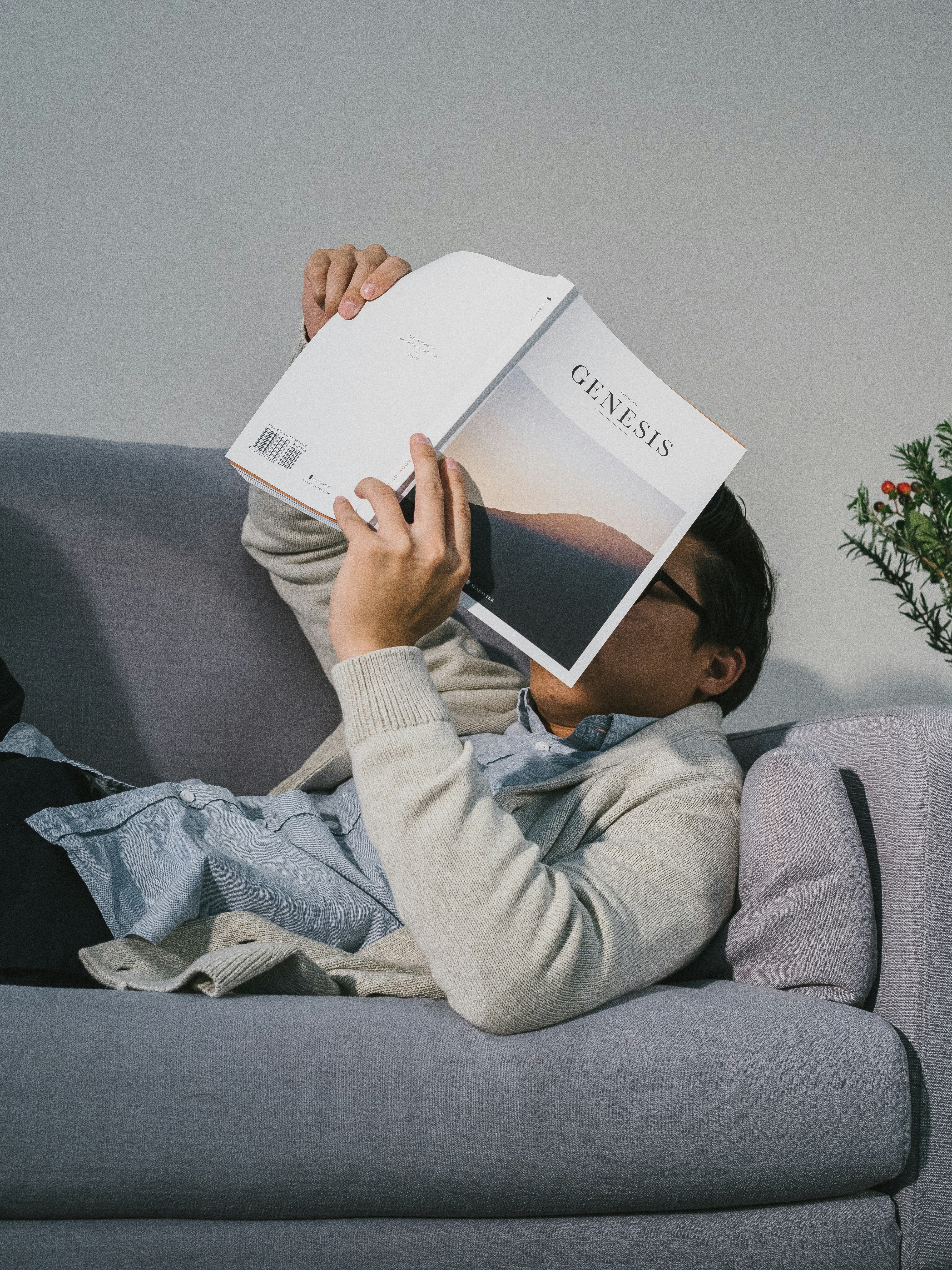 man reading book while lying on gray fabric sofa