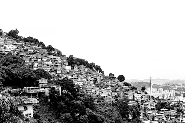 A densely populated hillside neighborhood with numerous similar-looking houses stacked closely together, surrounded by lush vegetation. The landscape extends to an urban area in the distance, featuring taller buildings and a broader cityscape.