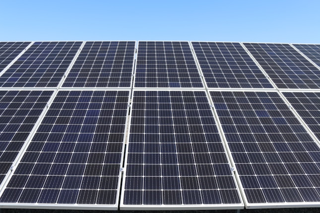 A field of solar panels is positioned under a clear blue sky. The panels are arranged in rows, capturing sunlight efficiently. The sleek, reflective surface of the panels gives a modern and clean look.