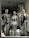 Three metallic mannequins are displayed in a store window, each dressed in distinctive swimwear. The mannequins have a shiny, reflective surface, with two wearing black swimsuits and one in a silver outfit. The setting includes floral arrangements in the background, creating an elegant atmosphere.