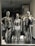 Three metallic mannequins are displayed in a store window, each dressed in distinctive swimwear. The mannequins have a shiny, reflective surface, with two wearing black swimsuits and one in a silver outfit. The setting includes floral arrangements in the background, creating an elegant atmosphere.