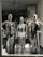 Three metallic mannequins are displayed in a store window, each dressed in distinctive swimwear. The mannequins have a shiny, reflective surface, with two wearing black swimsuits and one in a silver outfit. The setting includes floral arrangements in the background, creating an elegant atmosphere.