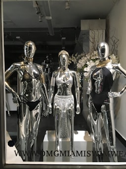 Three metallic mannequins are displayed in a store window, each dressed in distinctive swimwear. The mannequins have a shiny, reflective surface, with two wearing black swimsuits and one in a silver outfit. The setting includes floral arrangements in the background, creating an elegant atmosphere.