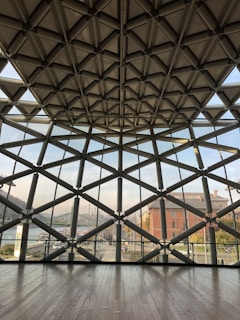 The interior view features a geometric, crisscrossing metal framework that forms a large window overlooking a cityscape. Beyond the window, a bridge spans a body of water, and brick buildings and trees are visible. The floor is made of wood, adding warmth to the otherwise industrial aesthetic.