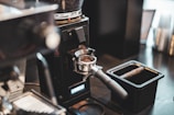 A close-up view of a coffee grinder dispensing freshly ground coffee into a portafilter in a cafe setting. The coffee beans are visible in the hopper at the top, while the ground coffee is poured into the handle below. The scene includes a black container and blurred background indicating a busy countertop.