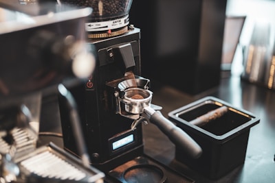 A close-up view of a coffee grinder dispensing freshly ground coffee into a portafilter in a cafe setting. The coffee beans are visible in the hopper at the top, while the ground coffee is poured into the handle below. The scene includes a black container and blurred background indicating a busy countertop.