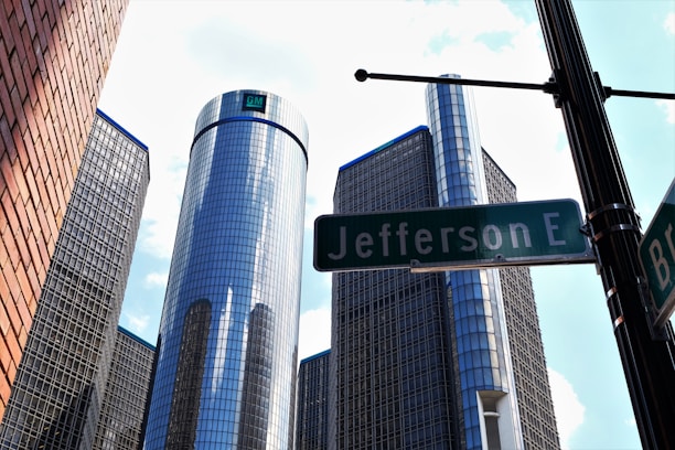 Several high-rise buildings with reflective glass facades dominate the skyline, capturing blue and cloudy sky reflections. A street sign marked 'Jefferson E' is prominently foregrounded, with a brick wall to the left.