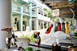 A collection of colorful metal animal sculptures including a swan with a large beak, a donkey, and a brightly patterned owl are displayed on a bench. In the background, there are mannequins dressed in vibrant clothes outside an art gallery. The setting appears to be an outdoor market or art space with a rustic wooden ceiling and pastel-colored walls.