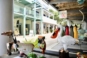 A collection of colorful metal animal sculptures including a swan with a large beak, a donkey, and a brightly patterned owl are displayed on a bench. In the background, there are mannequins dressed in vibrant clothes outside an art gallery. The setting appears to be an outdoor market or art space with a rustic wooden ceiling and pastel-colored walls.