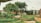 A lush garden setting featuring a variety of green plants, trees, and shrubs. Large boulders are scattered around, and a red brick pathway curves through the scene. A metal lamppost stands on the right, and rows of tall shrubs line the background alongside a metal structure.