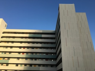 A multi-storey building with a modern architectural design features clean, straight lines and rectangular patterns. The facade displays a mix of narrow, vertical colored strips, mostly muted tones with a few pops of red. The structure's side is bathed in warm sunlight with a clear blue sky providing a serene backdrop.