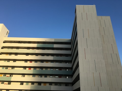 A multi-storey building with a modern architectural design features clean, straight lines and rectangular patterns. The facade displays a mix of narrow, vertical colored strips, mostly muted tones with a few pops of red. The structure's side is bathed in warm sunlight with a clear blue sky providing a serene backdrop.