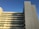 A multi-storey building with a modern architectural design features clean, straight lines and rectangular patterns. The facade displays a mix of narrow, vertical colored strips, mostly muted tones with a few pops of red. The structure's side is bathed in warm sunlight with a clear blue sky providing a serene backdrop.