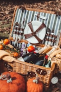 A wicker picnic basket filled with various items including a plate secured with leather straps, cutlery, and bottles of champagne and water. The basket is surrounded by fallen autumn leaves and pumpkins, and includes apples and snacks.