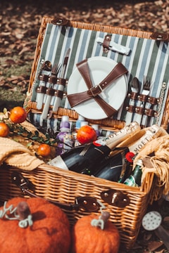 A wicker picnic basket filled with various items including a plate secured with leather straps, cutlery, and bottles of champagne and water. The basket is surrounded by fallen autumn leaves and pumpkins, and includes apples and snacks.