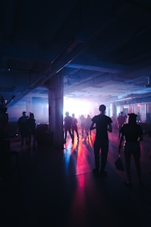 Silhouetted figures of people dancing in a dimly lit space with colorful lights casting vibrant reflections on the floor. The room has an industrial feel with exposed beams and overhead pipes.