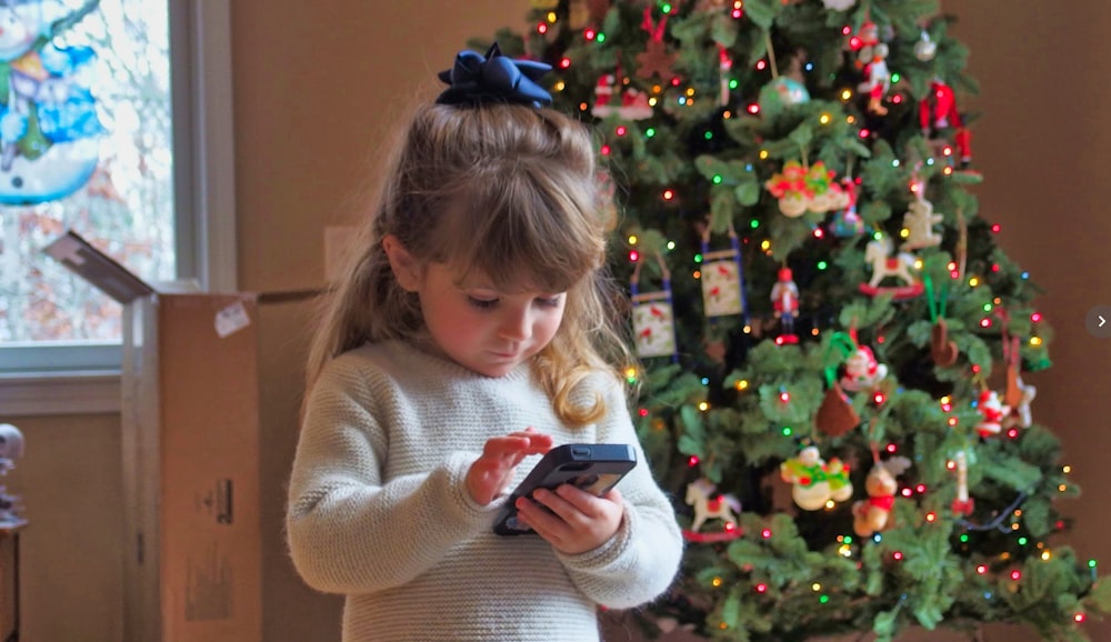 chica sosteniendo un teléfono inteligente cerca del árbol de Navidad