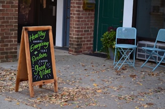 A sidewalk scene featuring a wooden sandwich board with colorful chalk writing, advertising healthy shakes, teas, and coffee with motivational phrases. Two light blue folding chairs are placed beside a window with a potted plant next to them. The setting includes a brick wall and a green door, with scattered leaves on the ground.