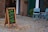 A sidewalk scene featuring a wooden sandwich board with colorful chalk writing, advertising healthy shakes, teas, and coffee with motivational phrases. Two light blue folding chairs are placed beside a window with a potted plant next to them. The setting includes a brick wall and a green door, with scattered leaves on the ground.