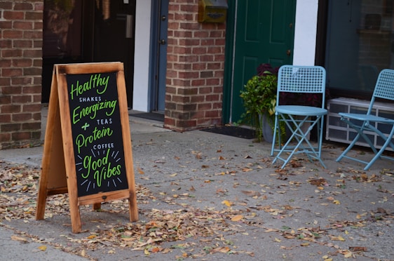 A sidewalk scene featuring a wooden sandwich board with colorful chalk writing, advertising healthy shakes, teas, and coffee with motivational phrases. Two light blue folding chairs are placed beside a window with a potted plant next to them. The setting includes a brick wall and a green door, with scattered leaves on the ground.