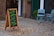 A sidewalk scene featuring a wooden sandwich board with colorful chalk writing, advertising healthy shakes, teas, and coffee with motivational phrases. Two light blue folding chairs are placed beside a window with a potted plant next to them. The setting includes a brick wall and a green door, with scattered leaves on the ground.