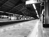 A black and white image of a modern train station platform, featuring high ceilings with metal beams and multiple signs in both English and Hindi providing directions. The platform is empty except for a few people in the distance. Fluorescent lights line the ceiling, casting reflections on the polished floor.
