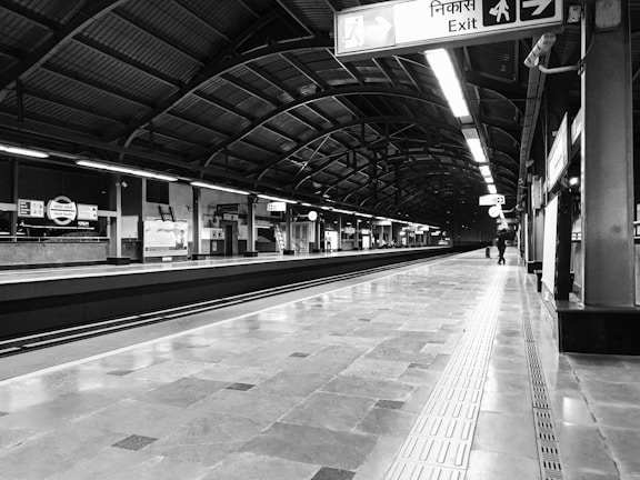 A black and white image of a modern train station platform, featuring high ceilings with metal beams and multiple signs in both English and Hindi providing directions. The platform is empty except for a few people in the distance. Fluorescent lights line the ceiling, casting reflections on the polished floor.