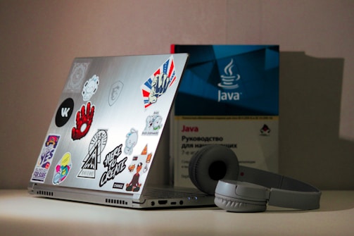 A laptop covered with various stickers is placed on a surface. Next to it is a book about Java programming, partially visible in the background. A pair of headphones rests beside the laptop, which has a metallic finish.