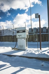 A diesel fuel pump stands amidst a snowy environment, with a background featuring a high fence and industrial structures. The ground is covered with thick, untouched snow, and the sky is partly cloudy with patches of bright blue visible. The light casts distinct shadows, suggesting a sunny yet cold day.