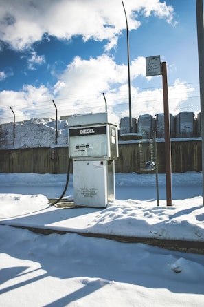 A diesel fuel pump stands amidst a snowy environment, with a background featuring a high fence and industrial structures. The ground is covered with thick, untouched snow, and the sky is partly cloudy with patches of bright blue visible. The light casts distinct shadows, suggesting a sunny yet cold day.