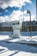 A diesel fuel pump stands amidst a snowy environment, with a background featuring a high fence and industrial structures. The ground is covered with thick, untouched snow, and the sky is partly cloudy with patches of bright blue visible. The light casts distinct shadows, suggesting a sunny yet cold day.