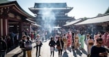 A bustling crowd gathers at a historic temple with traditional architecture. Many visitors are wearing colorful kimonos, while the air is filled with a light haze from burning incense. The setting is vibrant and lively, with a mix of tourists and locals.