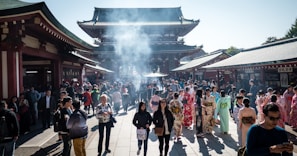 A bustling crowd gathers at a historic temple with traditional architecture. Many visitors are wearing colorful kimonos, while the air is filled with a light haze from burning incense. The setting is vibrant and lively, with a mix of tourists and locals.