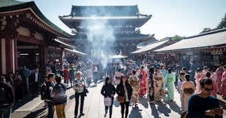 A bustling crowd gathers at a historic temple with traditional architecture. Many visitors are wearing colorful kimonos, while the air is filled with a light haze from burning incense. The setting is vibrant and lively, with a mix of tourists and locals.