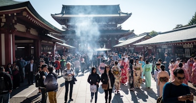 A bustling crowd gathers at a historic temple with traditional architecture. Many visitors are wearing colorful kimonos, while the air is filled with a light haze from burning incense. The setting is vibrant and lively, with a mix of tourists and locals.
