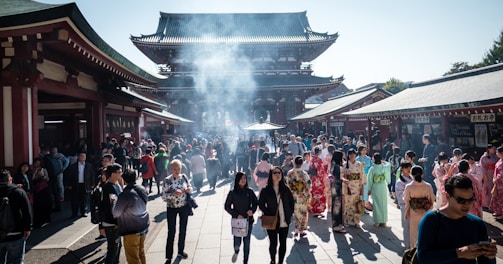 A bustling crowd gathers at a historic temple with traditional architecture. Many visitors are wearing colorful kimonos, while the air is filled with a light haze from burning incense. The setting is vibrant and lively, with a mix of tourists and locals.