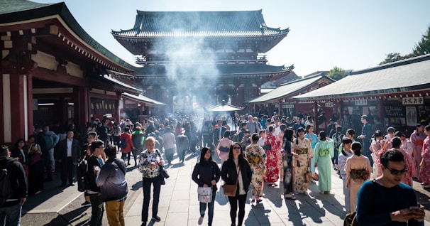 A bustling crowd gathers at a historic temple with traditional architecture. Many visitors are wearing colorful kimonos, while the air is filled with a light haze from burning incense. The setting is vibrant and lively, with a mix of tourists and locals.