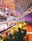A brightly lit shopping mall features a high, arched ceiling adorned with holiday decorations. The railing in the foreground is decorated with green garlands and golden ornaments. The vibrant purple and yellow lighting casts a warm glow over the space.