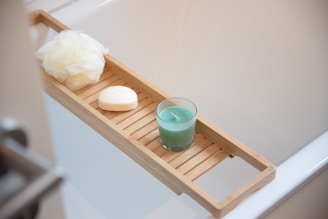 A wooden bath tray is positioned over a white bathtub. It holds a green scented candle, a white bar of soap with an embossed design, and an off-white bath sponge or loofah. The items suggest a serene and relaxing setting.