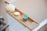 A wooden bath tray is positioned over a white bathtub. It holds a green scented candle, a white bar of soap with an embossed design, and an off-white bath sponge or loofah. The items suggest a serene and relaxing setting.