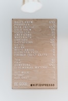 A well-organized menu board features various coffee and tea options with prices. The board has a perforated design, and prices are displayed next to each item. It includes drinks like daily brew, pourover, iced coffee, cold brew, espresso, and more. The bottom part displays a message encouraging kindness and the handle of a coffee shop.