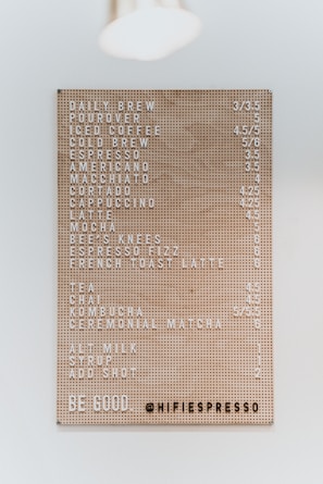 A well-organized menu board features various coffee and tea options with prices. The board has a perforated design, and prices are displayed next to each item. It includes drinks like daily brew, pourover, iced coffee, cold brew, espresso, and more. The bottom part displays a message encouraging kindness and the handle of a coffee shop.
