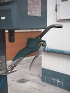 A close-up view of a green gas pump nozzle attached to a fuel dispenser. The background consists of a tiled wall and a concrete surface, revealing part of an industrial or service station environment.