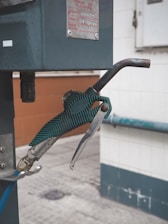 A close-up view of a green gas pump nozzle attached to a fuel dispenser. The background consists of a tiled wall and a concrete surface, revealing part of an industrial or service station environment.