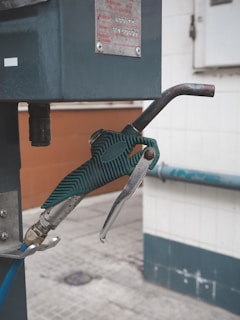 A close-up view of a green gas pump nozzle attached to a fuel dispenser. The background consists of a tiled wall and a concrete surface, revealing part of an industrial or service station environment.