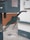 A close-up view of a green gas pump nozzle attached to a fuel dispenser. The background consists of a tiled wall and a concrete surface, revealing part of an industrial or service station environment.